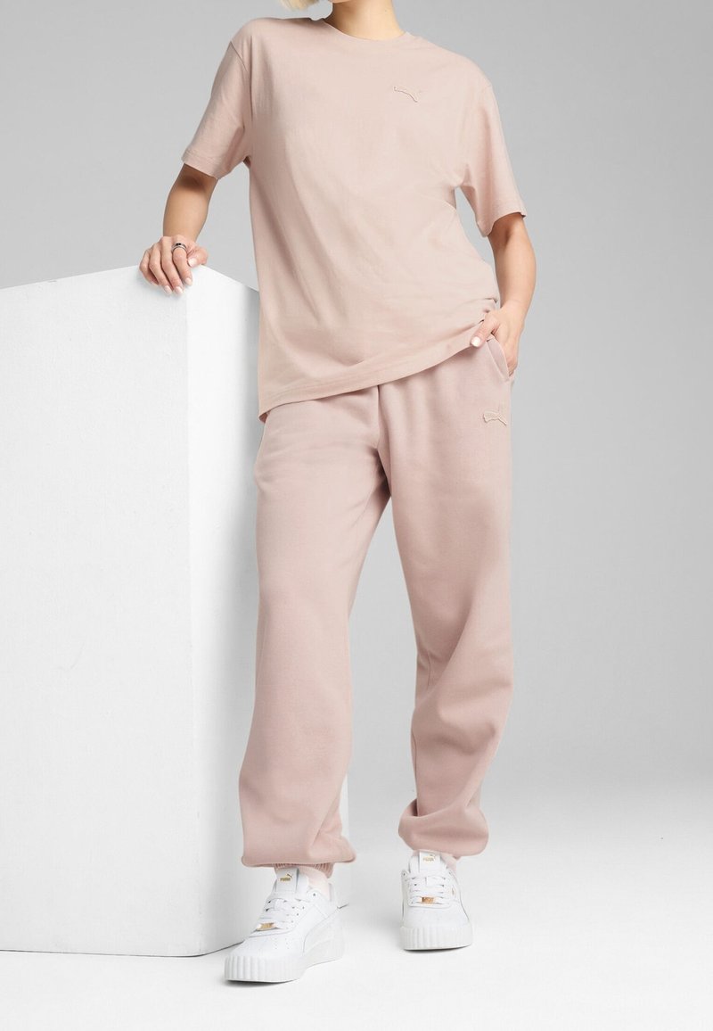 Personne portant un t-shirt rose pâle Puma et un pantalon de survêtement assorti avec des baskets blanches Puma, debout et appuyée contre un bloc blanc.