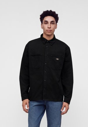 Ung mand med krøllet hår iført en sort skjorte med knapper og brystlommer samt blå jeans, står foran en ensfarvet lys baggrund.