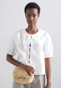 Bluză albă cu mâneci bufante, guler rotund și detaliu legat în față. Geantă încrucișată bej, țesută, cu închidere, având modele texturate.