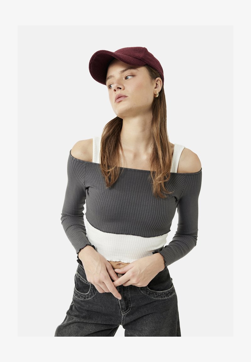 Joven con gorra burdeos, top corto gris con hombros descubiertos sobre camiseta blanca sin mangas y vaqueros negros, posando con las manos cerca de la cintura.