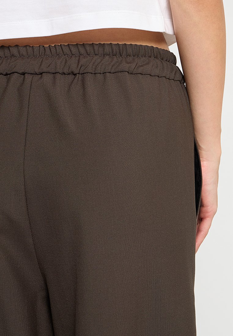 Braune, elastische Hose mit glatter Textur und lockerer Passform. Verfügt über eine Seitentasche und wird von hinten mit einem kurzen Oberteil darüber gezeigt.