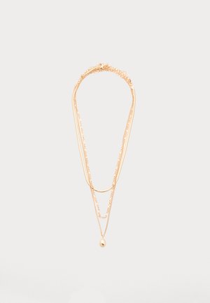 Trīsslāņu zelta kaklarota ar plānām ķēdītēm, izliekta stienīša rotājums un maza ovāla piekariņa garākajā ķēdītē, attēlota uz balta fona.