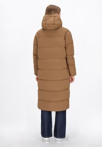 Geacă puffer maro cu glugă detașabilă, cu matlasare orizontală, mâneci lungi și un design de lungime medie, combinată cu blugi închiși la culoare și pantofi.