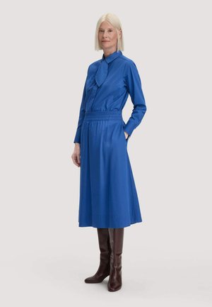 Frau mit glatten blonden Haaren, die ein langärmeliges blaues Kleid und dunkelbraune kniehohe Stiefel trägt und mit einer Hand in der Tasche steht.