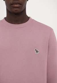Homme portant un sweat-shirt mauve à col rond avec un petit emblème de cheval rayé multicolore sur le côté gauche de la poitrine.