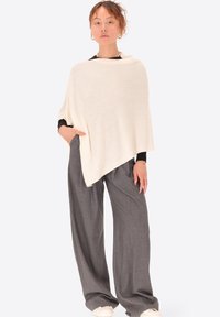 Poncho color crema realizzato in tessuto morbido, che cade morbido con un orlo irregolare, abbinato a pantaloni grigi a gamba larga e sneakers bianche.