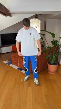 Vit bomullst-shirt med en liten lila logotyp, kombinerad med blå byxor med gula detaljer och olika logotyper. Silverfärgade skor.