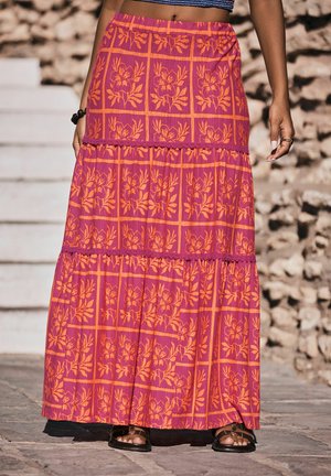 Femme portant une longue jupe rose avec un motif quadrillé floral orange, associée à des sandales noires, debout sur un pavé en pierre près des marches.