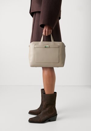 Persona con abrigo marrón oscuro y botas a juego, sosteniendo un bolso rectangular beige con asas en la parte superior y bolsillo frontal.