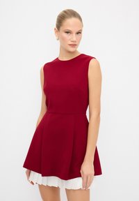 Robe sans manches rouge avec un corsage ajusté, une jupe évasée et une couche inférieure plissée blanche superposée. Tissu lisse et décolleté arrondi.
