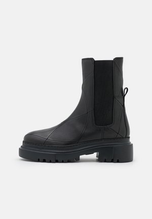 Bota de tobillo de cuero negro con suela gruesa y con tacos, panel lateral elástico y lengüeta en el talón, mostrada de perfil sobre fondo blanco.