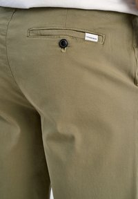 Khakigrüne Hose aus Baumwollstoff. Besitzt eine Gesäßtasche mit einem schwarzen Knopf und ein Etikett mit der Aufschrift "LINDGEBRGH." Glatte Textur.