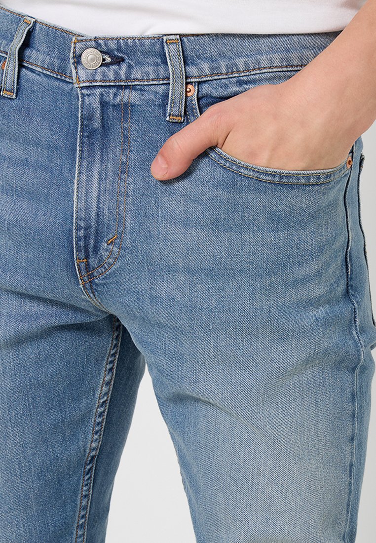 Jean en denim bleu clair avec un design classique à cinq poches, des coutures contrastantes et un bouton en métal visible à la taille.