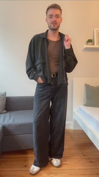 Melns zip-up jaka pār muguru brūnu topu, apvienota ar vaļīgām melnām biksēm. Baltas sneakers ar pelēkajiem un melnajiem akcentiem. Koka grīda un gaiša fons.