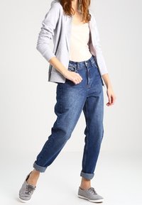 Grauer Kapuzenpullover mit Reißverschluss über einem hellbeigen Oberteil, kombiniert mit hoch taillierten, locker sitzenden blauen Jeans, die an den Bündchen umgekrempelt sind, und grauen Turnschuhen.