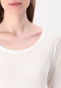 Top in maglia a coste color crema con scollatura ampia e arrotondata, caratterizzato da una texture sottile, maniche lunghe e una silhouette aderente.