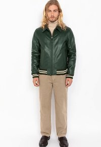 Veste bombardier en cuir vert avec une fermeture éclair à l'avant, des poignets et un col côtelés. Portée avec un pantalon beige et des chaussures foncées, posant devant un fond blanc.