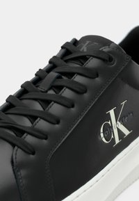 Zwarte leren sneakers met ronde neuzen, platte veters en een witte rubberen zool, met een subtiel "CK"-logo aan de zijkant. Gladde textuur.