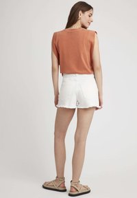 Haut de tank top court couleur rouille avec des manches courtes, assorti à un short en denim blanc découpé et des sandales à lanières beige. Les ourlets effilochés sont visibles.