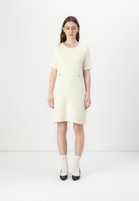 Robe tissée crème à manches courtes, surface texturée, deux petites poches avant avec des accents dorés, associée à des chaussettes blanches et des chaussures à talons noires.