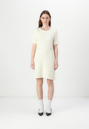 Woven cream dress with short sleeves, textured surface, two small front pockets with gold accents, paired with white socks and black heeled shoes.
