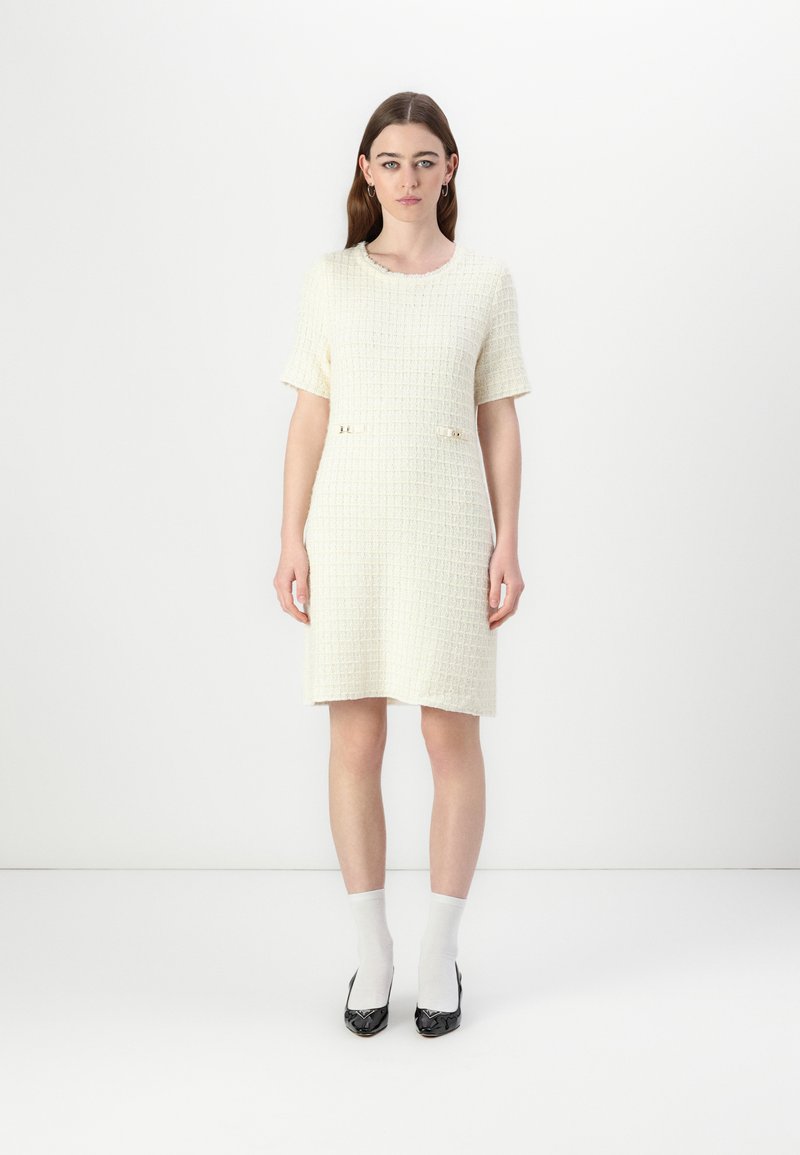 Robe tissée crème à manches courtes, surface texturée, deux petites poches avant avec des accents dorés, associée à des chaussettes blanches et des chaussures à talons noires.