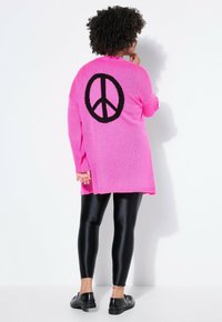 Jasně růžový pletený svetr s velkým černým znakem míru na zádech, kombinovaný s lesklými černými legínami a černými botami.