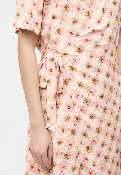 Κοντινό πλάνο στο μπράτσο και το χέρι ενός ατόμου που φοράει ένα ανοιχτό ροζ φόρεμα τύπου wrap με ροζ και κίτρινο λουλουδένιο σχέδιο και ζώνη από υφασμα.