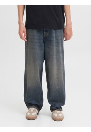 Personne portant un jean bleu délavé et ample ainsi que des baskets beiges, debout devant un fond clair uni.
