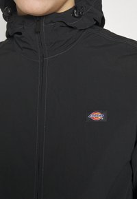 Svart huvtröja med en slät yta, med dragkedja framtill och en framträdande Dickies-logopatch på bröstet. Vita sömmar som accentuerar.
