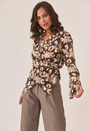 Blusa envolvente floral de tela oscura con patrones en beige y marrón, con mangas largas y un ajuste estructurado. Combinada con pantalones gris de corte ajustado.