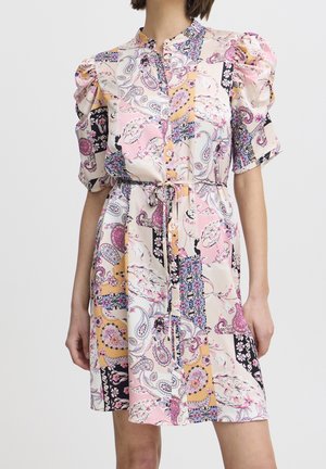 Kobieta ubrana w sukienkę o długości do kolan z kwiatowym wzorem w stylu patchwork, z krótkimi bufkowatymi rękawami i przewiązanym paskiem w talii, w pastelowych kolorach różu, żółci i czerni.