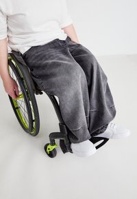 Sive hlače iz denima z ohlapnim krojem in subtilnimi poškodovanji, v kombinaciji z belimi športnimi copati, nosi oseba, ki sedi v invalidskem vozičku z zelenimi detajli.