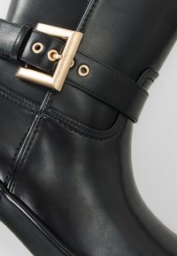 Cizmă din piele neagră cu detalii cusute, având o cataramă pătrată aurie, gravată cu "Armani Exchange", pe brățara de la gleznă.