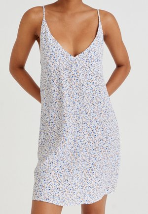 Femme portant une robe sans manches à col en V avec de fines bretelles et un petit imprimé floral bleu sur fond blanc, mains derrière le dos.