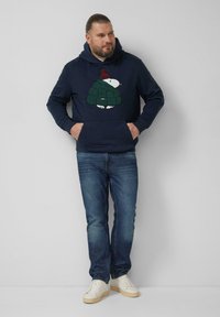 Granatowa bluza z kapturem z dzierganym motywem żółwia w czerwonym kapeluszu, zestawiona z niebieskimi dżinsami i białymi trampkami. Klasyczny, casualowy wygląd.