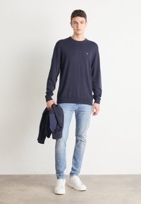 Tamsiai mėlynas ilgomis rankovėmis megztinis, su raukiniais ant kaklo ir rankogalių, derinamas su šviesiai mėlynomis džinsomis ir baltais sportbačiais, laikydamas atitinkamą striukę.