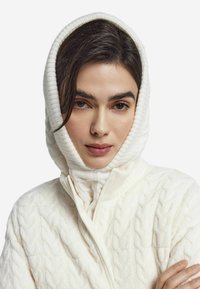 Biały sweter z kapturem w stylu z warkoczami, z wysokim kołnierzem i zintegrowanym kapturem. Miękka faktura i zapięcie na zamek błyskawiczny. Wzór prezentuje wypukłe, przeplatane warkocze.