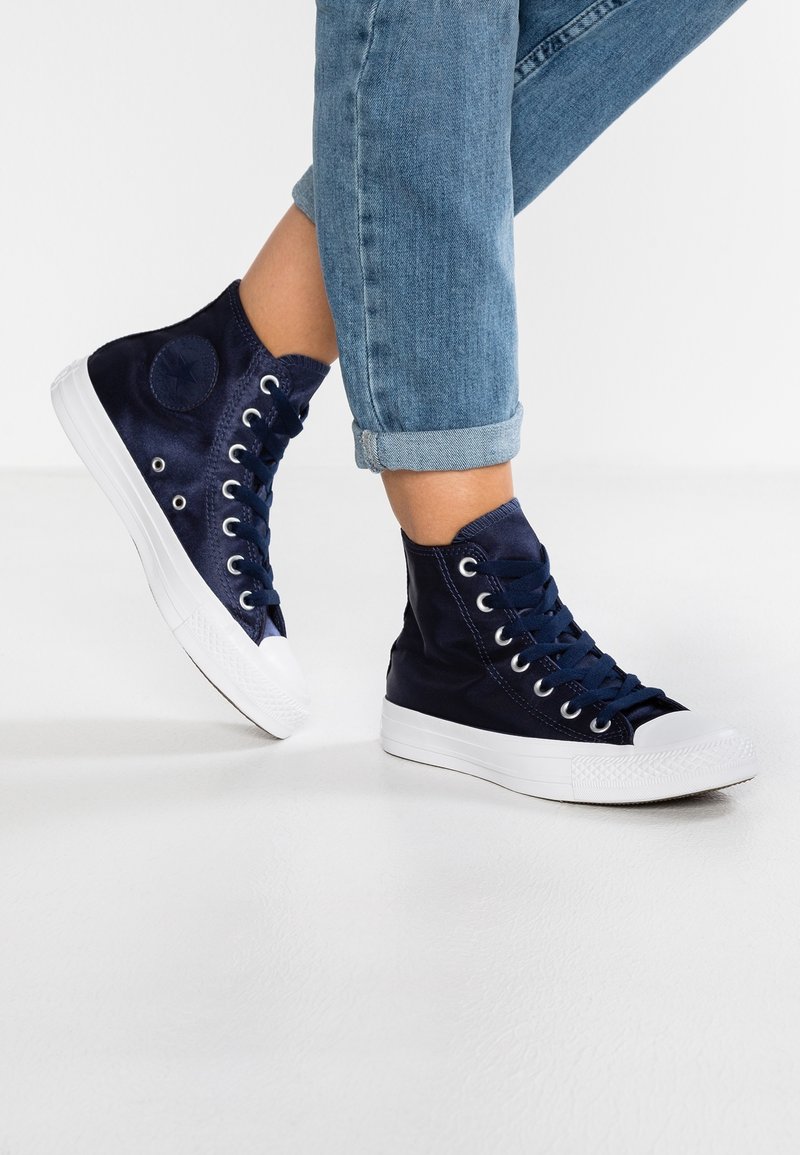 Personne portant un jean bleu retroussé et des baskets montantes bleu marine debout sur un sol blanc.