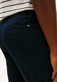 Gros plan d'une personne portant un pantalon bleu marine foncé avec une poche arrière boutonnée et un petit logo rouge, blanc et bleu près de la poche.