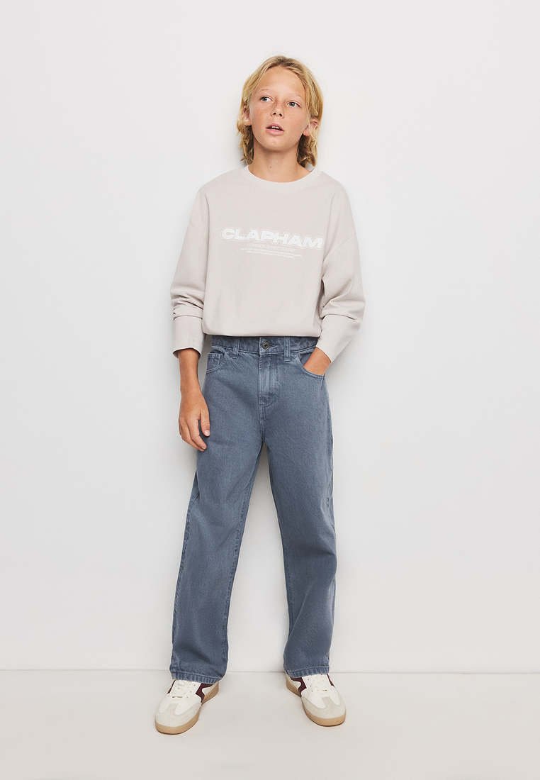 Enfant blond debout contre un mur blanc portant un sweat beige avec le texte "CLAPHAM", un jean bleu et des baskets blanches.