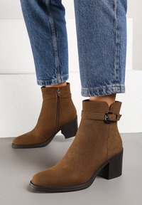 Bottines en suede marron, avec un bout pointu, un talon bloc, une fermeture éclair sur le côté, et une sangle avec une boucle. Portées avec un jean en denim raccourci.