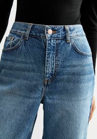 High-waisted blue jeans with a classic fit, featuring subtle fading, orange stitching, and front pockets with a metal button closure.