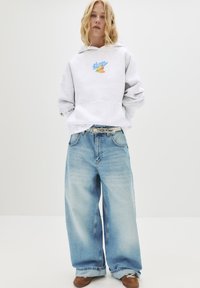 Persona con capelli biondi indossa una felpa grigia chiara con grafica, pantaloni a gamba larga blu sbiaditi con una cintura bianca e scarpe marroni, in piedi.