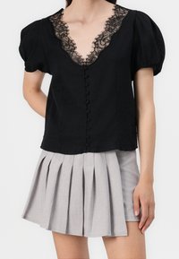 Blouse noire avec un col en dentelle et des manches bouffantes, présentant une rangée de boutons. Associée à une jupe plissée gris clair.