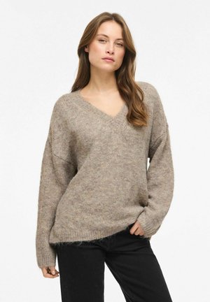 Kvinde med langt brunt hår iført en løs, beige strikket V-hals trøje og sorte bukser, stående med den ene hånd i lommen mod hvid baggrund.