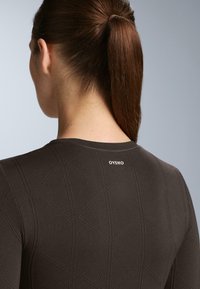 Žena s hnedými vlasmi v cope, oblečená v tmavom štruktúrovanom dlhom rukáve s logom "OYSHO" na hornej časti chrbta, na jednolatom šedom pozadí.