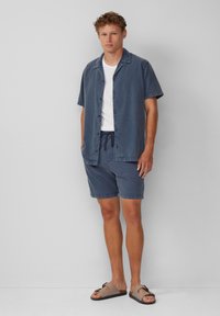 Herren Kurzarm-Jeanshemd in Dunkelblau, kombiniert mit passenden Shorts und braunen Sandalen. Mit Knopfverschluss und einem Kordelzugbund versehen.