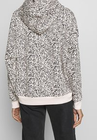 Person som bär en ljusfärgad huvtröja med svarta abstrakta svängda mönster och ljusa ärmslut, i kombination med mörka byxor, vänd bort.