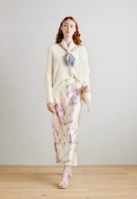 Suéter de punto crema con un corte holgado, falda midi floral en colores pasteles y sandalias; presenta una bufanda celeste atada alrededor del cuello.